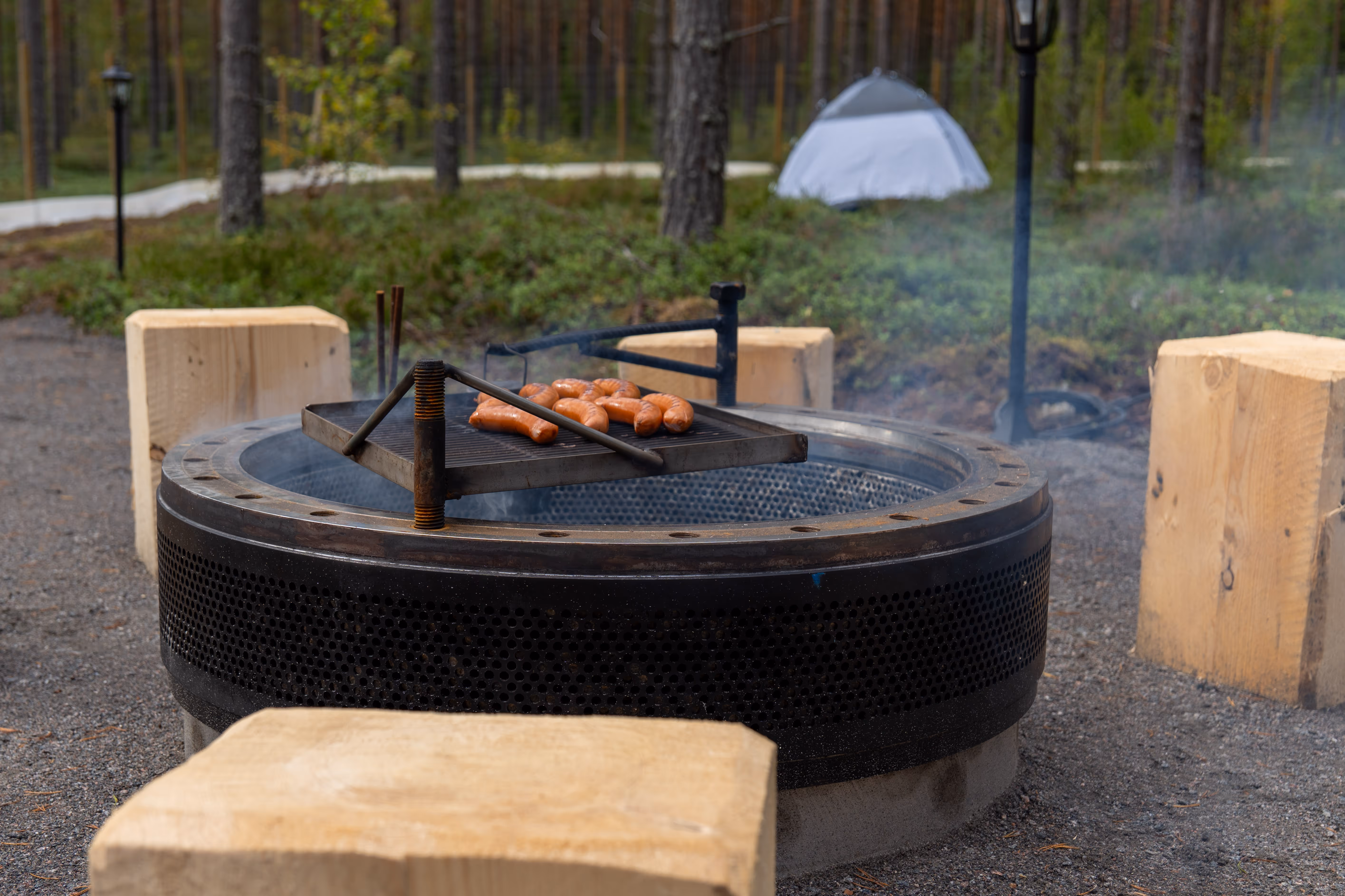 Korkeakosken koirametsän viihtyisä nuotiopaikka, jossa makkarat paistuvat metallisella ritilällä suuren, pyöreän tulisijan yllä. Ympärillä on puisia pölkkyjä istuimina.