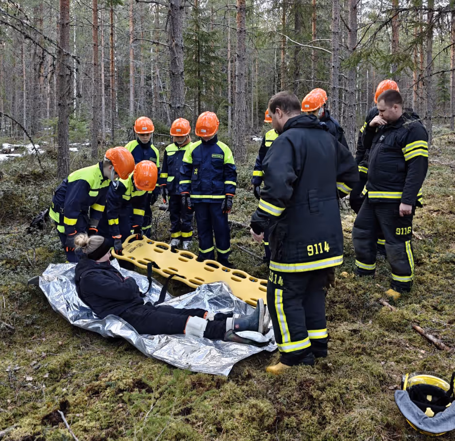 Palomiehet ja nuorisopalokuntalaiset ovat kokoontuneet loukkaantunutta esittävän henkilön ympärille pelastusharjoituksissa Korkeakosken koirametsässä. Henkilö makaa avaruuslakalla, ja vieressä on keltainen rankalauta.
